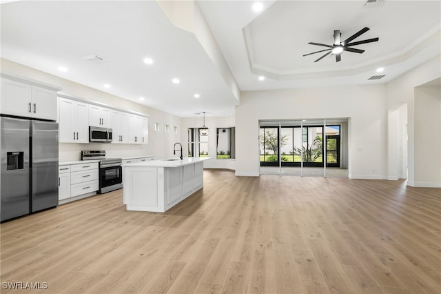 kitchen featuring appliances with stainless steel finishes, a tray ceiling, an island with sink, white cabinets, and light wood-type flooring