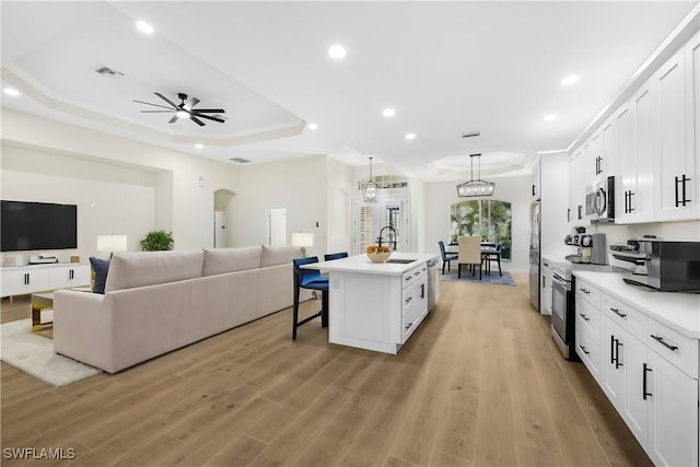 kitchen with appliances with stainless steel finishes, a tray ceiling, white cabinets, a center island with sink, and decorative light fixtures