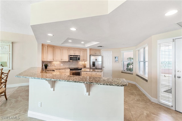 kitchen with light brown cabinetry, appliances with stainless steel finishes, a tray ceiling, kitchen peninsula, and a breakfast bar area