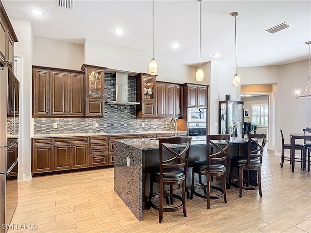 kitchen with a center island with sink, decorative light fixtures, wall chimney range hood, and light hardwood / wood-style flooring