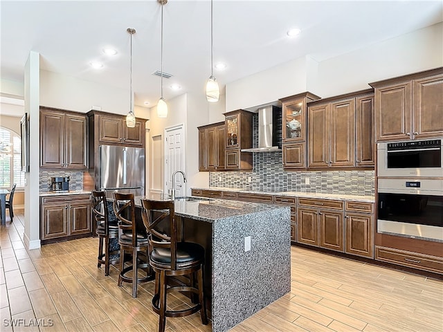 kitchen with a kitchen bar, wall chimney exhaust hood, sink, a center island with sink, and hanging light fixtures