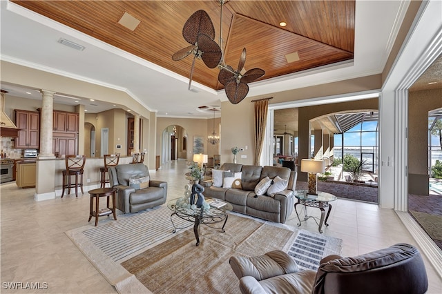 tiled living room with a raised ceiling, ceiling fan with notable chandelier, ornate columns, wood ceiling, and crown molding