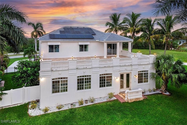 back of property at dusk with a lawn, a tile roof, fence, roof mounted solar panels, and stucco siding