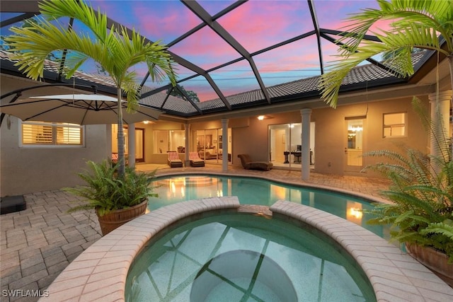 pool at dusk with an in ground hot tub, an outdoor hangout area, glass enclosure, and a patio area