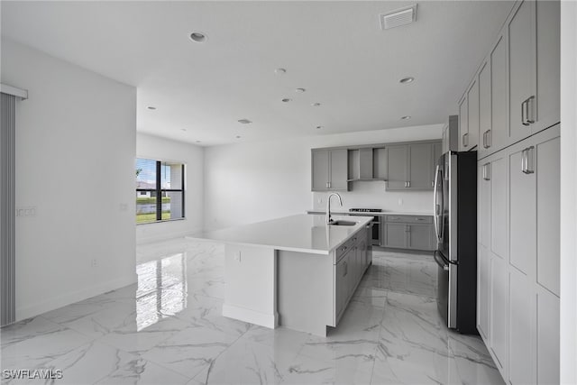 kitchen with sink, wall chimney range hood, gray cabinets, a center island with sink, and appliances with stainless steel finishes