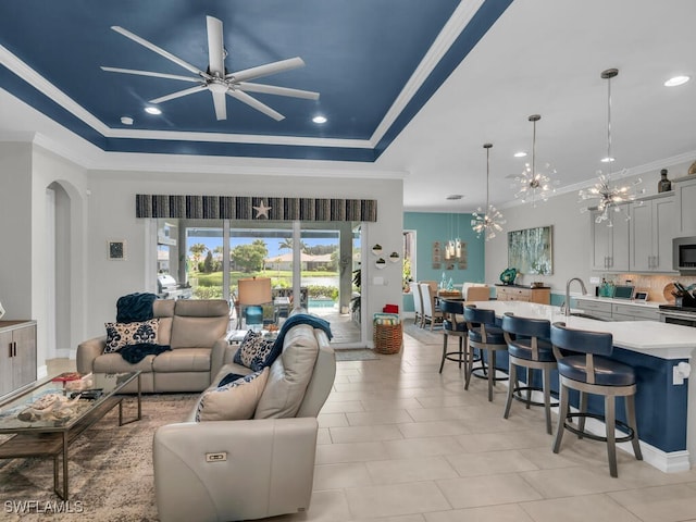 tiled living room with sink, ornamental molding, ceiling fan with notable chandelier, and a raised ceiling