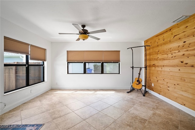 tiled empty room with a healthy amount of sunlight, ceiling fan, and wood walls