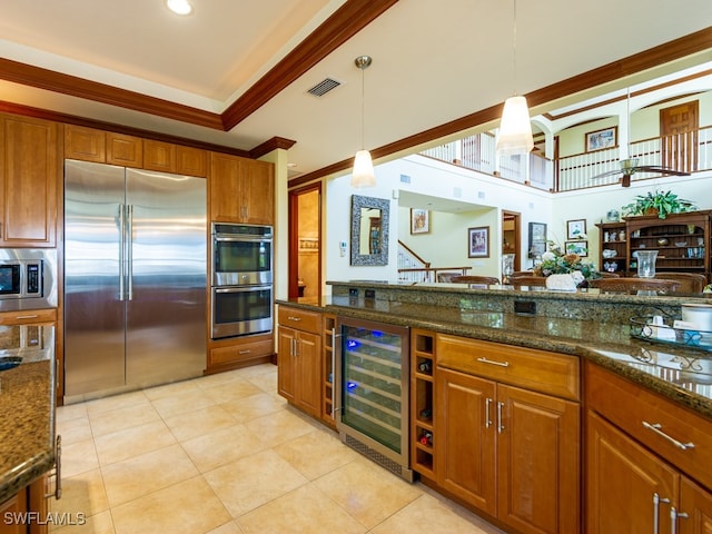 kitchen with hanging light fixtures, built in appliances, dark stone countertops, ornamental molding, and beverage cooler