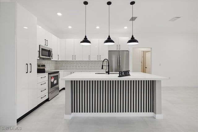 kitchen featuring a kitchen island with sink, white cabinetry, backsplash, stainless steel appliances, and decorative light fixtures