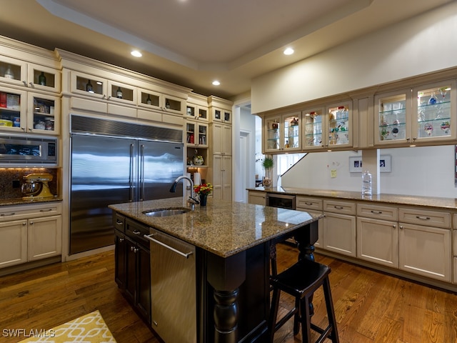 kitchen featuring light stone countertops, a kitchen island with sink, sink, built in appliances, and a breakfast bar area