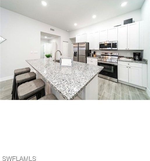 kitchen featuring light stone counters, a center island with sink, white cabinetry, appliances with stainless steel finishes, and a kitchen breakfast bar