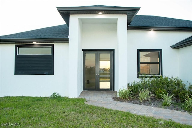 entrance to property with a shingled roof, french doors, a lawn, and stucco siding