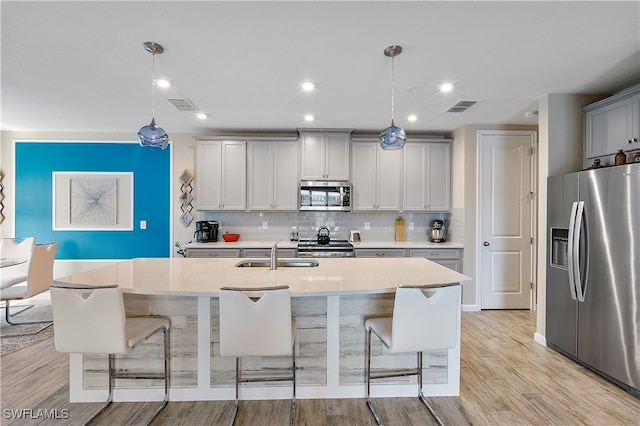 kitchen with a center island with sink, appliances with stainless steel finishes, decorative light fixtures, sink, and light hardwood / wood-style floors