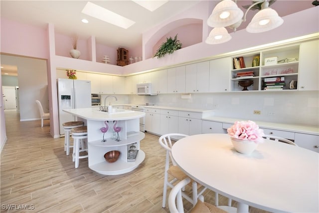 kitchen with a skylight, white appliances, decorative light fixtures, white cabinets, and an island with sink