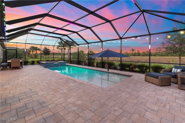 pool at dusk featuring glass enclosure, a patio area, and an in ground hot tub