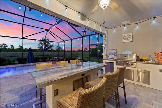 patio terrace at dusk with grilling area, an outdoor kitchen, sink, ceiling fan, and a lanai