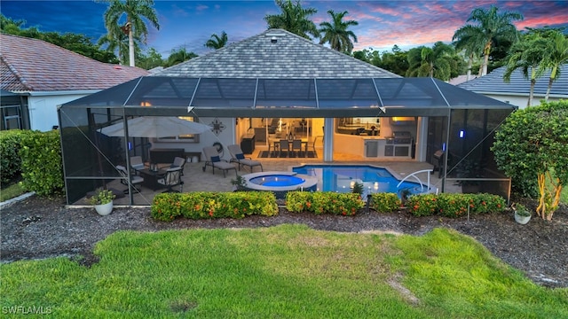 back house at dusk with a lanai, a patio area, a swimming pool with hot tub, and a lawn