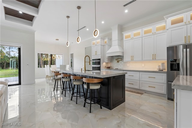 kitchen featuring premium range hood, decorative light fixtures, a center island with sink, white cabinetry, and stainless steel appliances