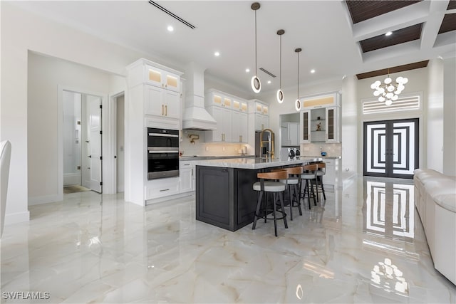 kitchen featuring white cabinets, decorative backsplash, a center island with sink, decorative light fixtures, and premium range hood