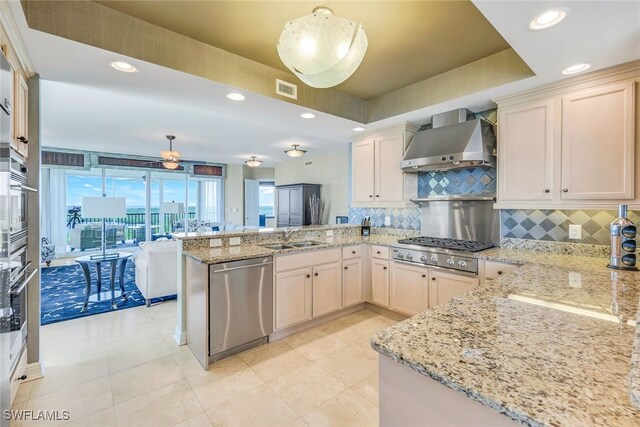 kitchen with tasteful backsplash, appliances with stainless steel finishes, hanging light fixtures, wall chimney exhaust hood, and light stone counters