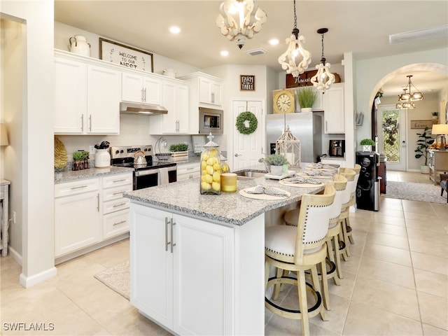 kitchen with light stone countertops, a kitchen island with sink, white cabinets, appliances with stainless steel finishes, and decorative light fixtures