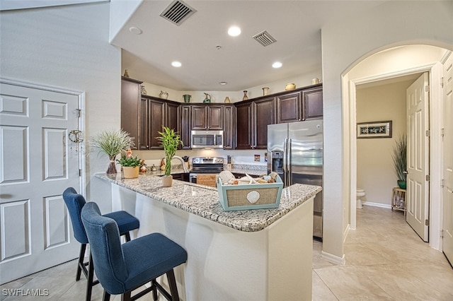 kitchen with light tile patterned floors, dark brown cabinetry, kitchen peninsula, light stone countertops, and appliances with stainless steel finishes