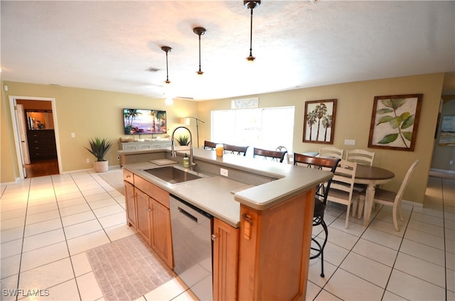 kitchen with an island with sink, light tile patterned floors, pendant lighting, stainless steel dishwasher, and sink