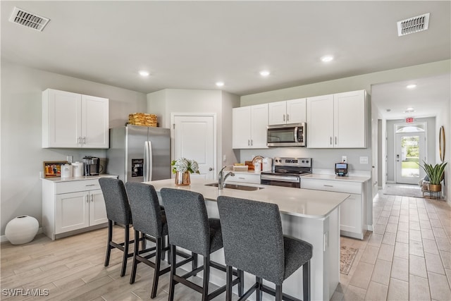 kitchen with sink, appliances with stainless steel finishes, a kitchen island with sink, white cabinets, and light wood-type flooring