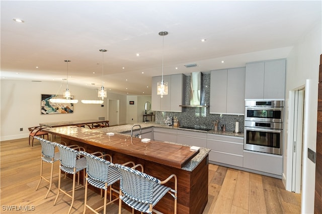 kitchen with light hardwood / wood-style flooring, stainless steel double oven, pendant lighting, and wall chimney range hood