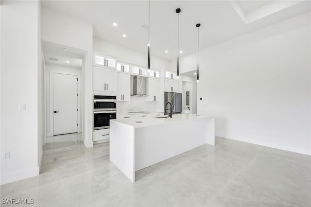 kitchen featuring an island with sink, wall chimney exhaust hood, glass insert cabinets, hanging light fixtures, and white cabinetry