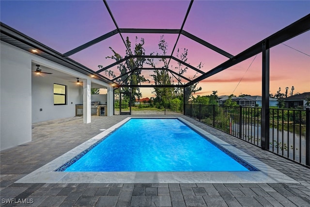 pool at dusk with a ceiling fan, area for grilling, glass enclosure, a water view, and a patio area