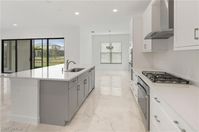 kitchen with wall chimney range hood, a kitchen island with sink, decorative light fixtures, sink, and white cabinetry