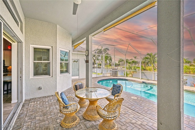 pool at dusk featuring glass enclosure, ceiling fan, and a patio