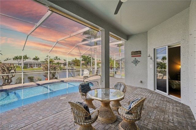 pool at dusk featuring a lanai, ceiling fan, a water view, and a patio