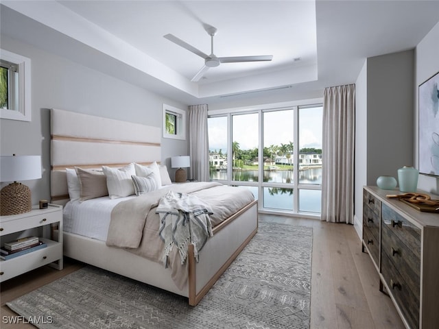 bedroom with access to outside, a raised ceiling, light hardwood / wood-style floors, ceiling fan, and a water view