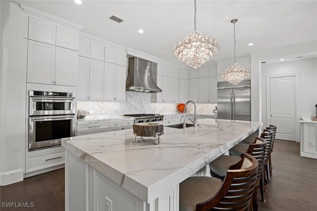 kitchen with wall chimney exhaust hood, dark hardwood / wood-style floors, a large island with sink, white cabinets, and appliances with stainless steel finishes