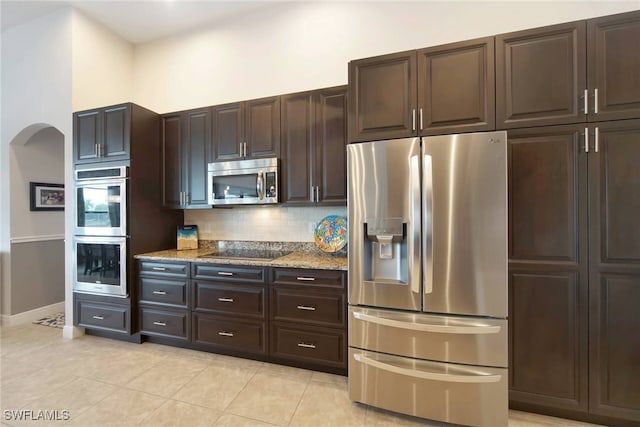 kitchen featuring light stone counters, light tile patterned flooring, dark brown cabinets, stainless steel appliances, and decorative backsplash