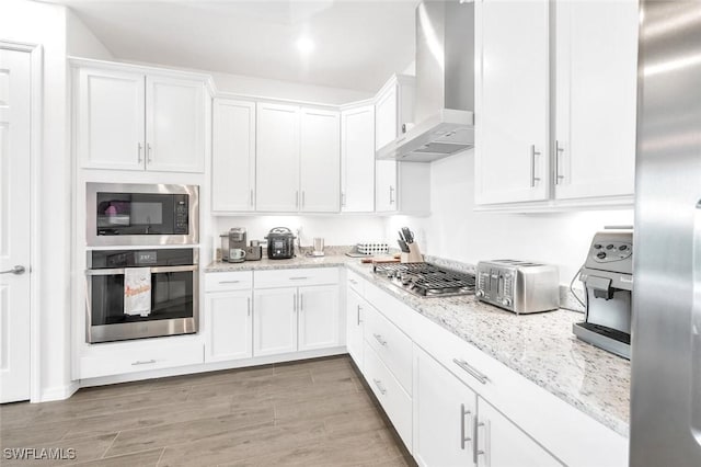 kitchen featuring stainless steel appliances, wall chimney range hood, white cabinetry, light stone countertops, and light wood-style floors