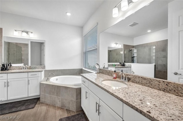 bathroom with a walk in shower, a bath, two vanities, wood finished floors, and recessed lighting