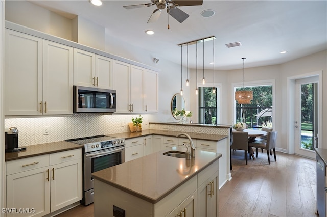 kitchen featuring kitchen peninsula, appliances with stainless steel finishes, a kitchen island with sink, sink, and pendant lighting