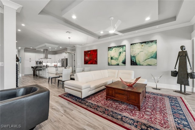 living room featuring light wood-type flooring, a tray ceiling, ceiling fan, and ornate columns