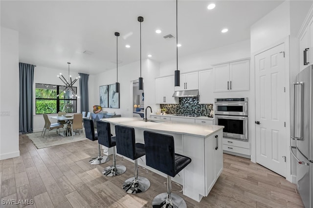 kitchen featuring stainless steel appliances, decorative light fixtures, white cabinetry, tasteful backsplash, and a center island with sink