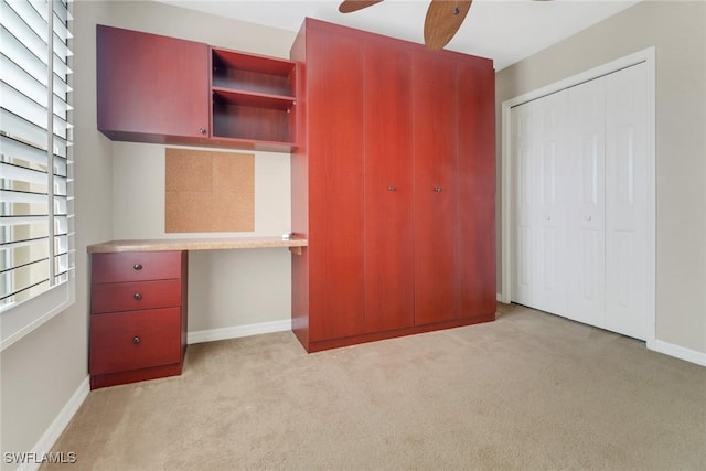 unfurnished bedroom with a closet, light colored carpet, built in study area, ceiling fan, and baseboards