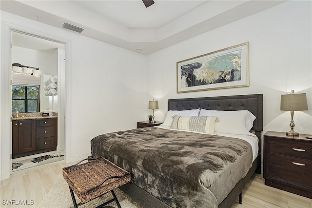 bedroom with light wood-type flooring, connected bathroom, a tray ceiling, and ceiling fan