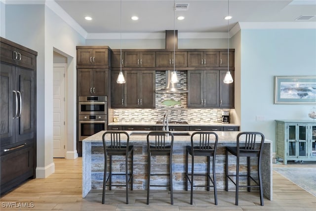 kitchen with wall chimney exhaust hood, a kitchen bar, a center island with sink, and double oven