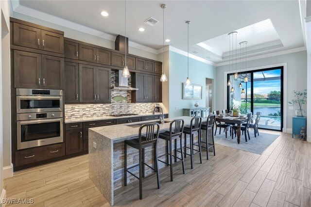 kitchen featuring stainless steel double oven, a tray ceiling, pendant lighting, a kitchen bar, and a center island with sink