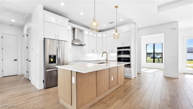 kitchen featuring stainless steel appliances, tasteful backsplash, light wood-style floors, wall chimney range hood, and an island with sink