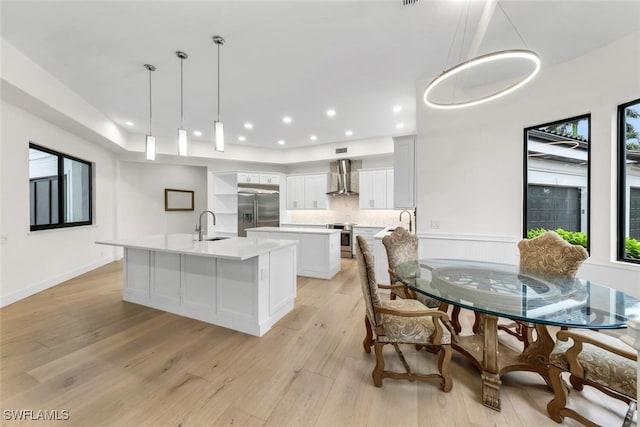 kitchen featuring white cabinetry, a kitchen island with sink, decorative light fixtures, appliances with stainless steel finishes, and wall chimney exhaust hood