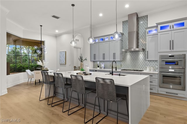 kitchen with sink, wall chimney exhaust hood, decorative light fixtures, a center island with sink, and light wood-type flooring