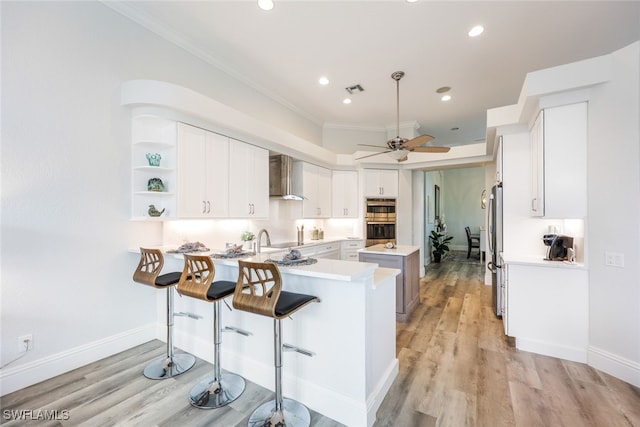 kitchen featuring wall chimney range hood, light hardwood / wood-style flooring, kitchen peninsula, crown molding, and white cabinetry